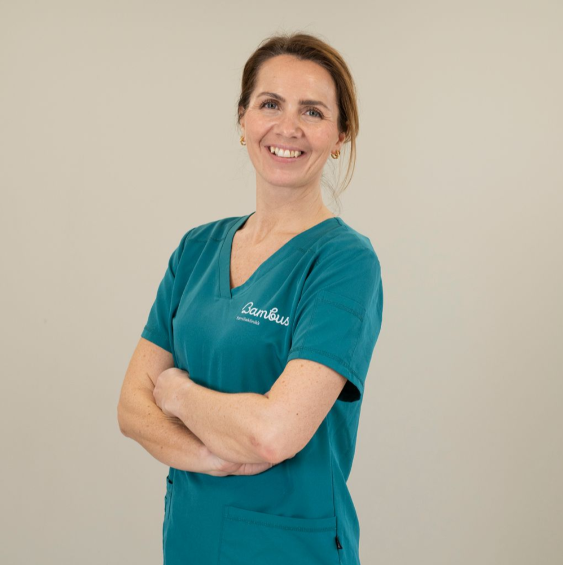 En smilende profesjonell i blågrønn skrubb med armene i kors, stående mot en nøytral bakgrunn.