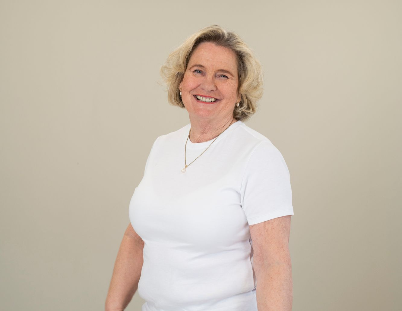 En smilende person med skulderlangt blondt hår iført en hvit V-hals-T-skjorte og et halskjede mot en enkel bakgrunn.