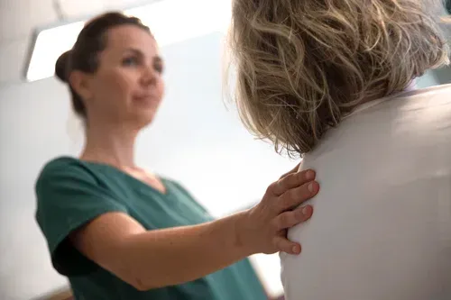 En helsepersonell i en grønn topp legger en hånd på en pasients skulder i en klinisk setting.