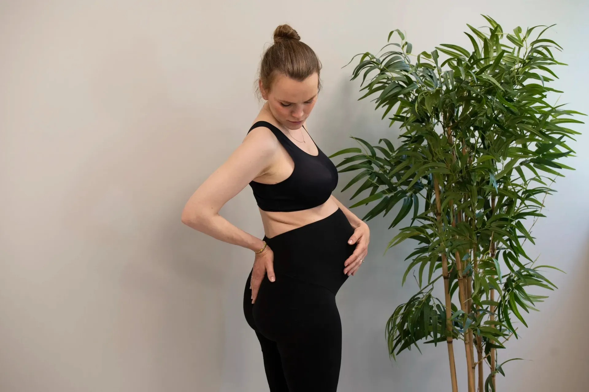 En gravid person iført svart sports-BH og leggings, står i profil med én hånd på ryggen og én på magen.