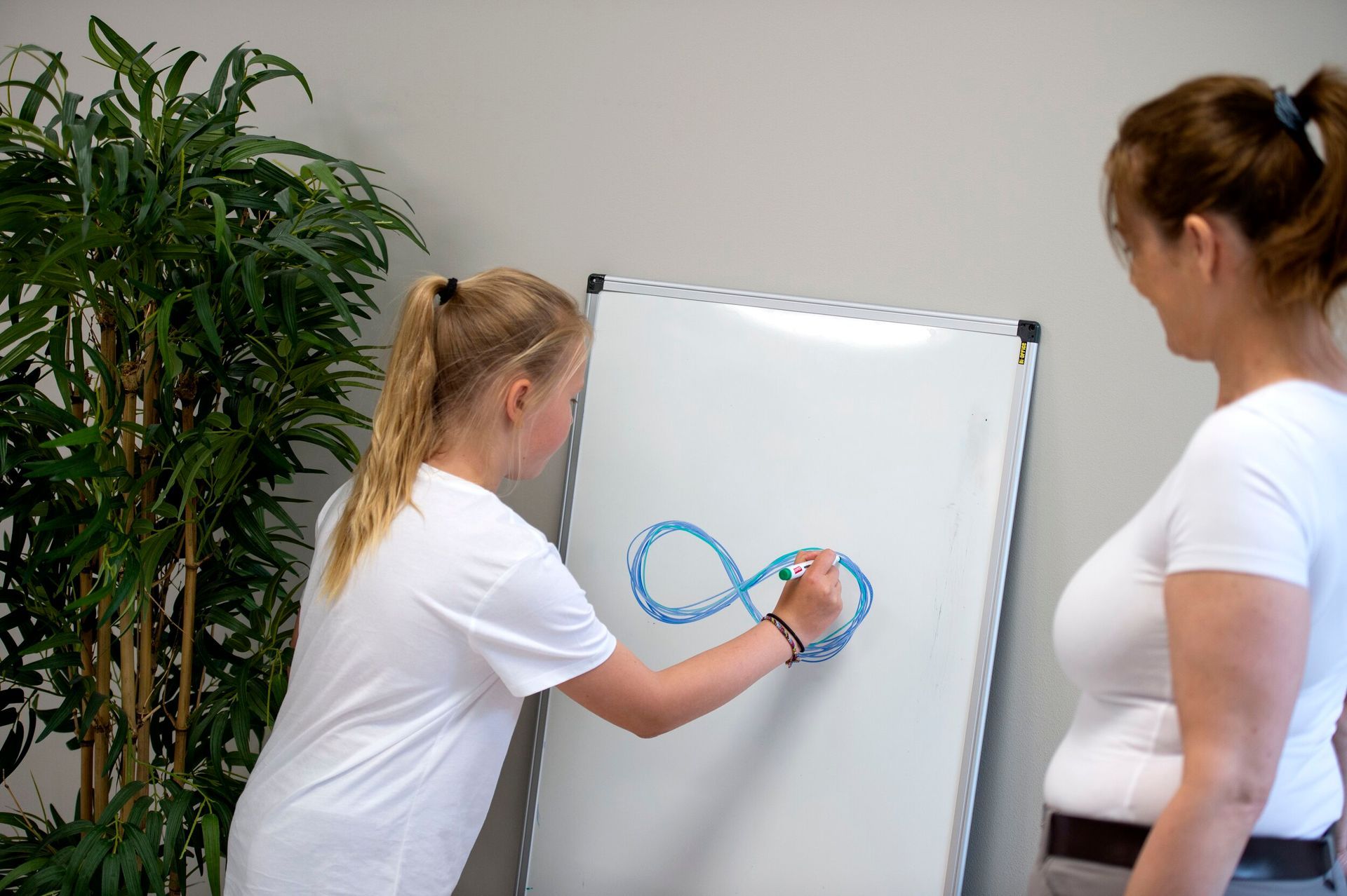 En person tegner et blått evighetssymbol på en tavle mens en annen person ser på fra siden.