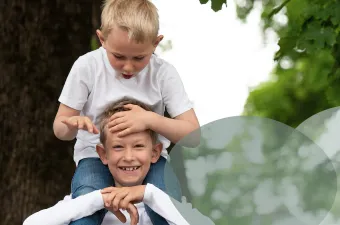 To personer i hvite skjorter utenfor; den ene sitter på den andres skuldre og smiler mens personen ovenfor berører håret deres.