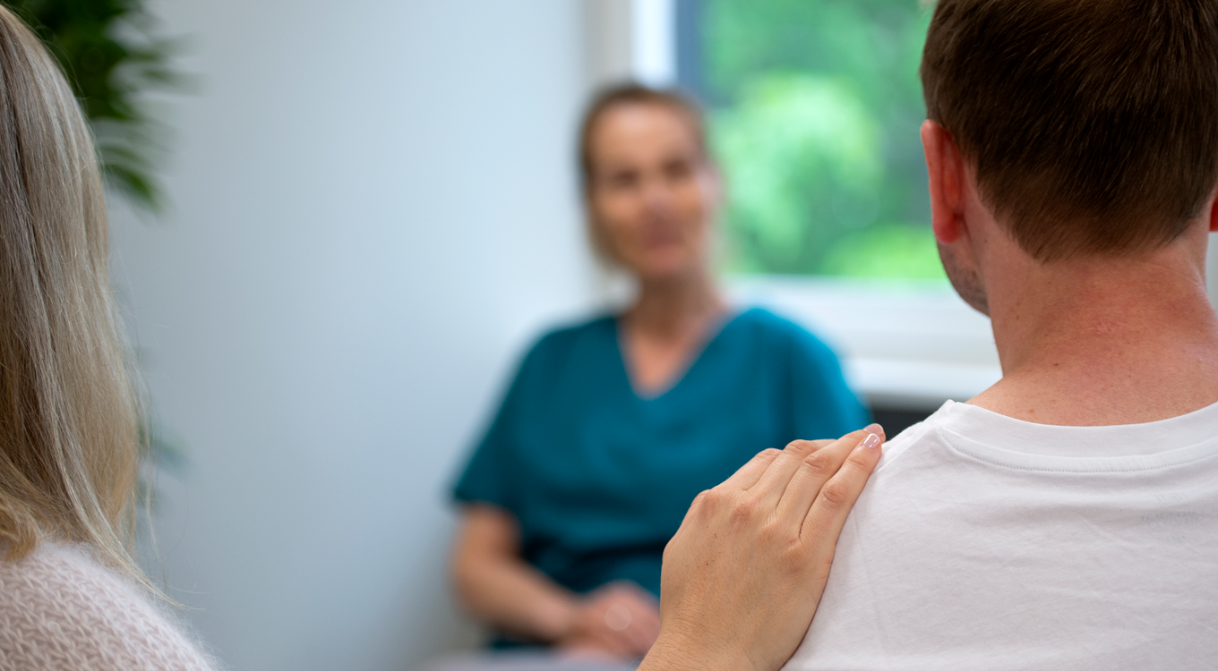 En person legger en trøstende hånd på en annens skulder i et uklart klinisk eller kontormiljø med en fagperson i nærheten.