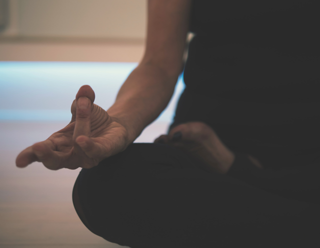 En person sitter i en meditativ positur med hånden i en mudra-gest mot en uskarp, lys bakgrunn.