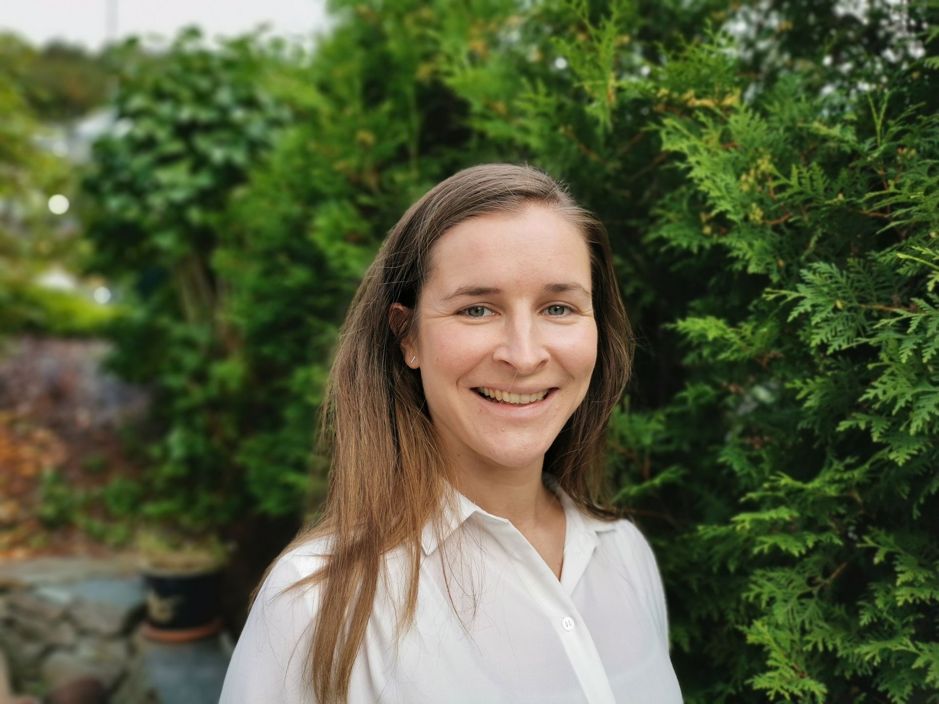 En smilende person med langt lysebrunt hår iført en hvit skjorte med krage, står utendørs foran grønne busker.