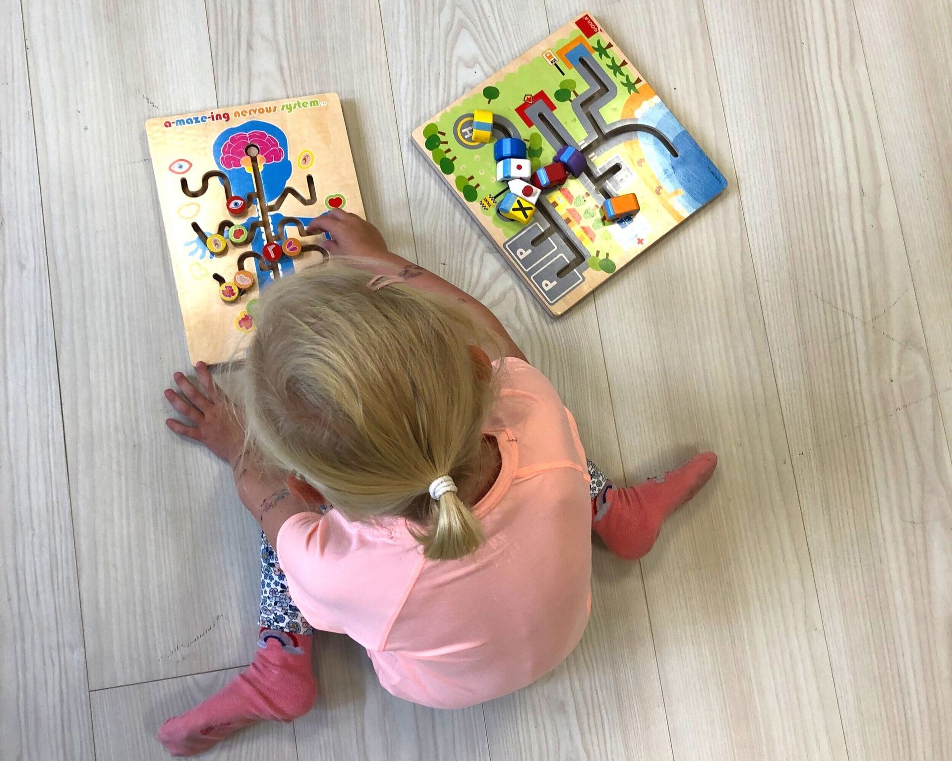 Et barn i en rosa skjorte sitter på et lyst tregulv og leker med to labyrintpuslespill i tre.