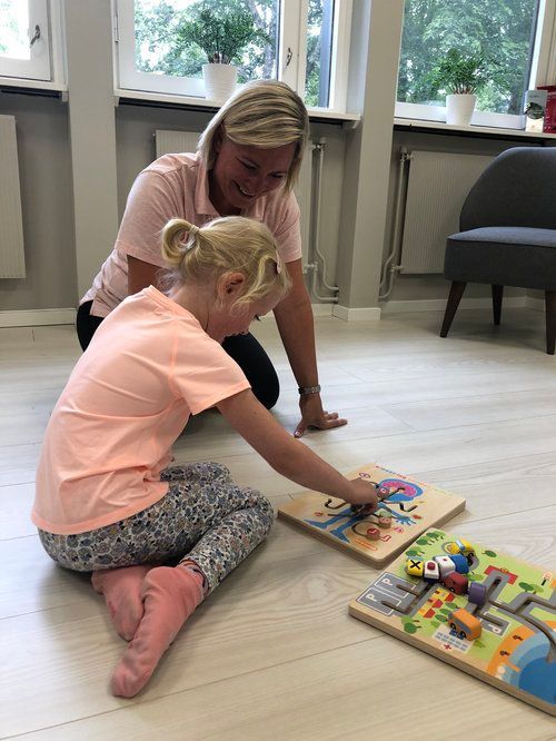 En person samhandler med et lite barn som leker med trebrettpuslespill på et lyst gulv.