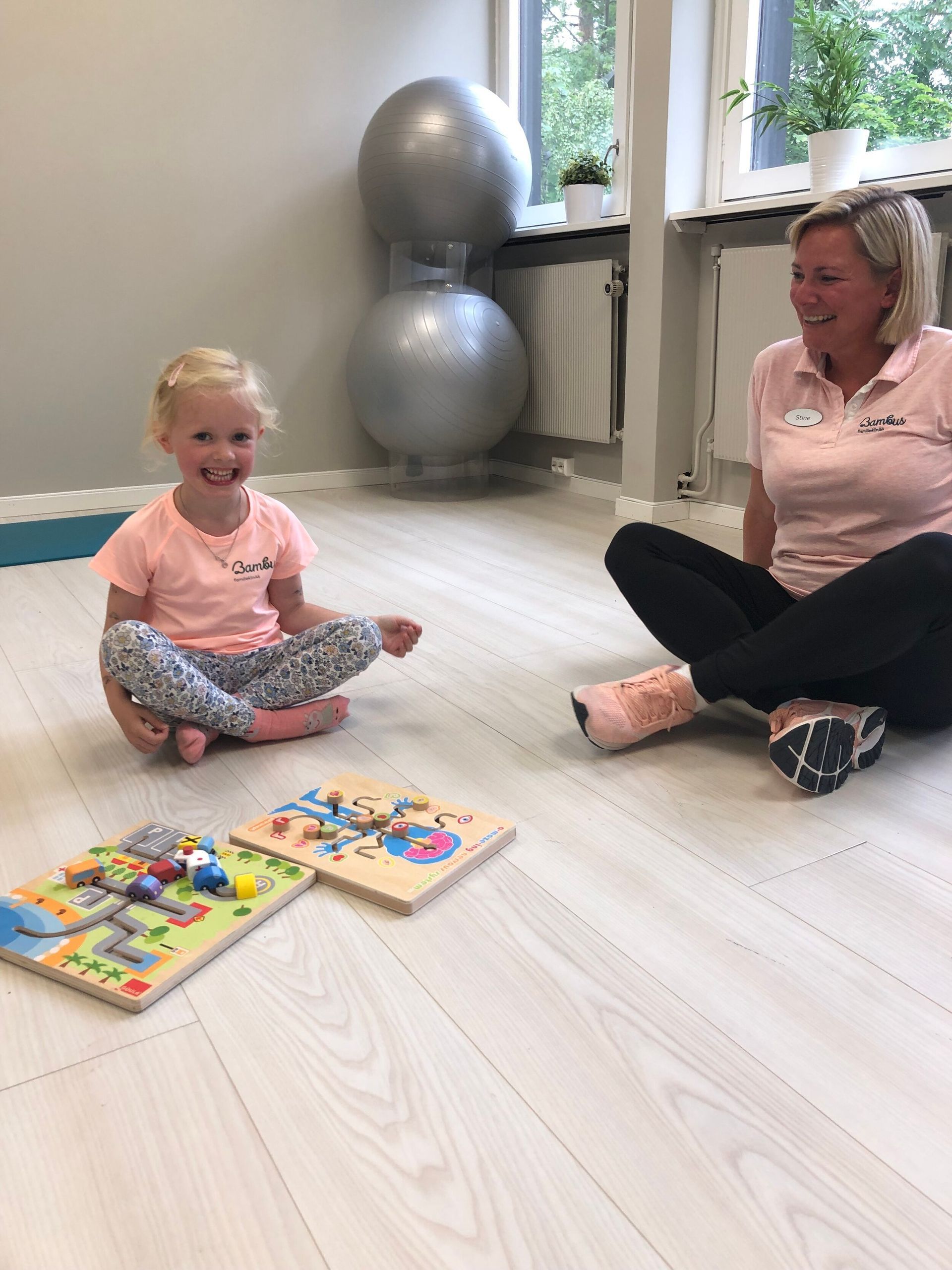 En smilende voksen og et barn sitter med beina i kors på et gulv og leker med trepuslespillbrett i et sterkt opplyst rom.