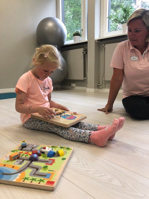 En person som leker med et trepuslespill mens han sitter på gulvet med en annen person som observerer.