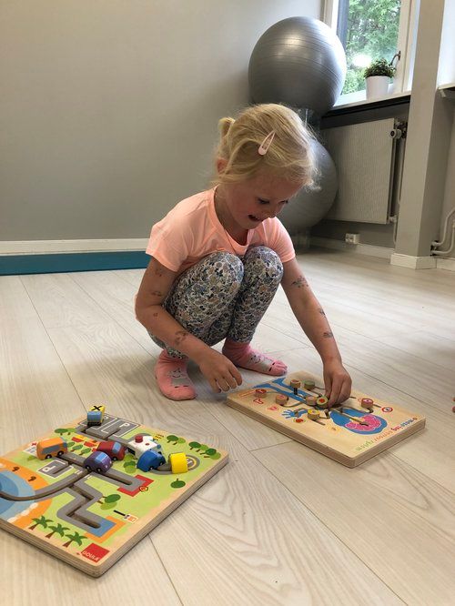 En person som kneler på et gulv leker med to trepuslespill, ett med en vei og det andre med fargerike figurer.