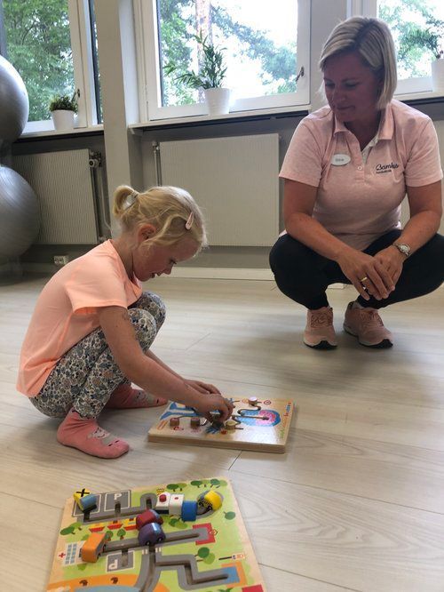 En person holder oppsyn med et barn som sitter på gulvet og er fokusert på å løse trepuslespill i et lyst innendørsrom.