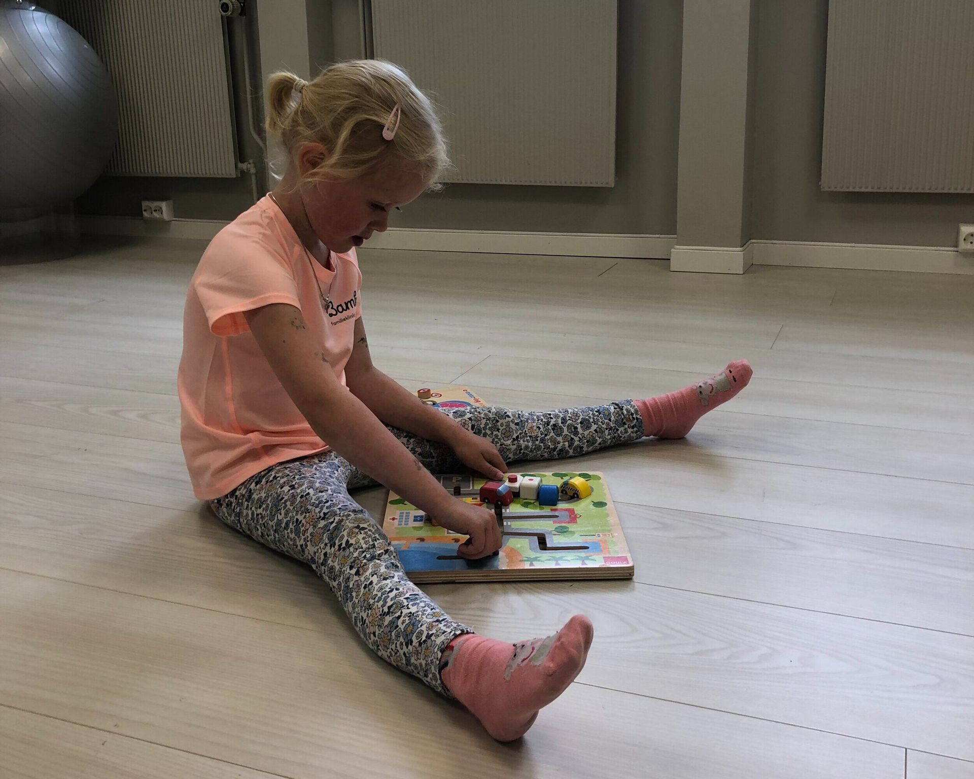 Et barn i en ferskenfarget skjorte og mønstrede leggings sitter på et lyst gulv og leker med et puslespillbrett i tre.