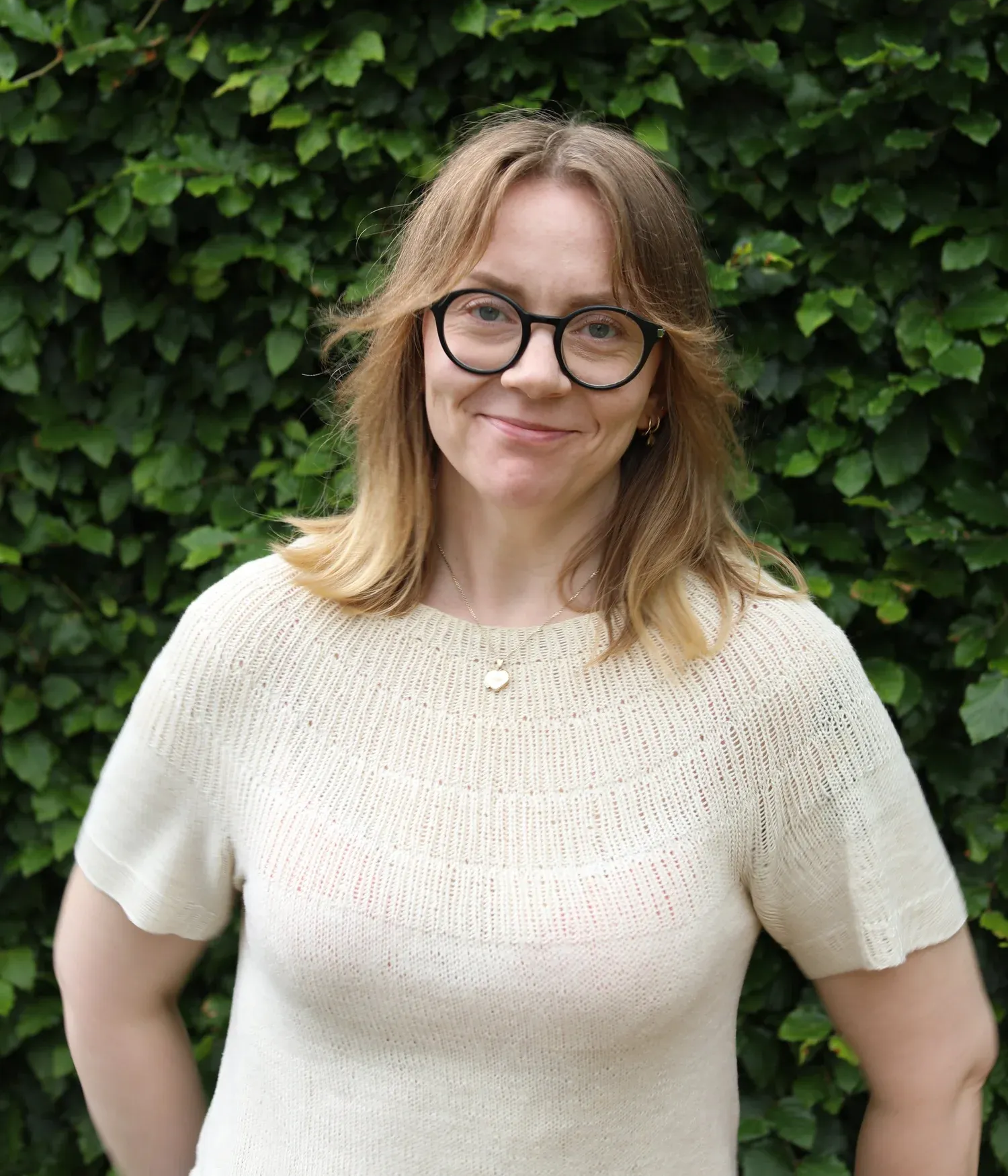 En person med lysebrunt hår og briller smiler foran en frodig grønn hekk.