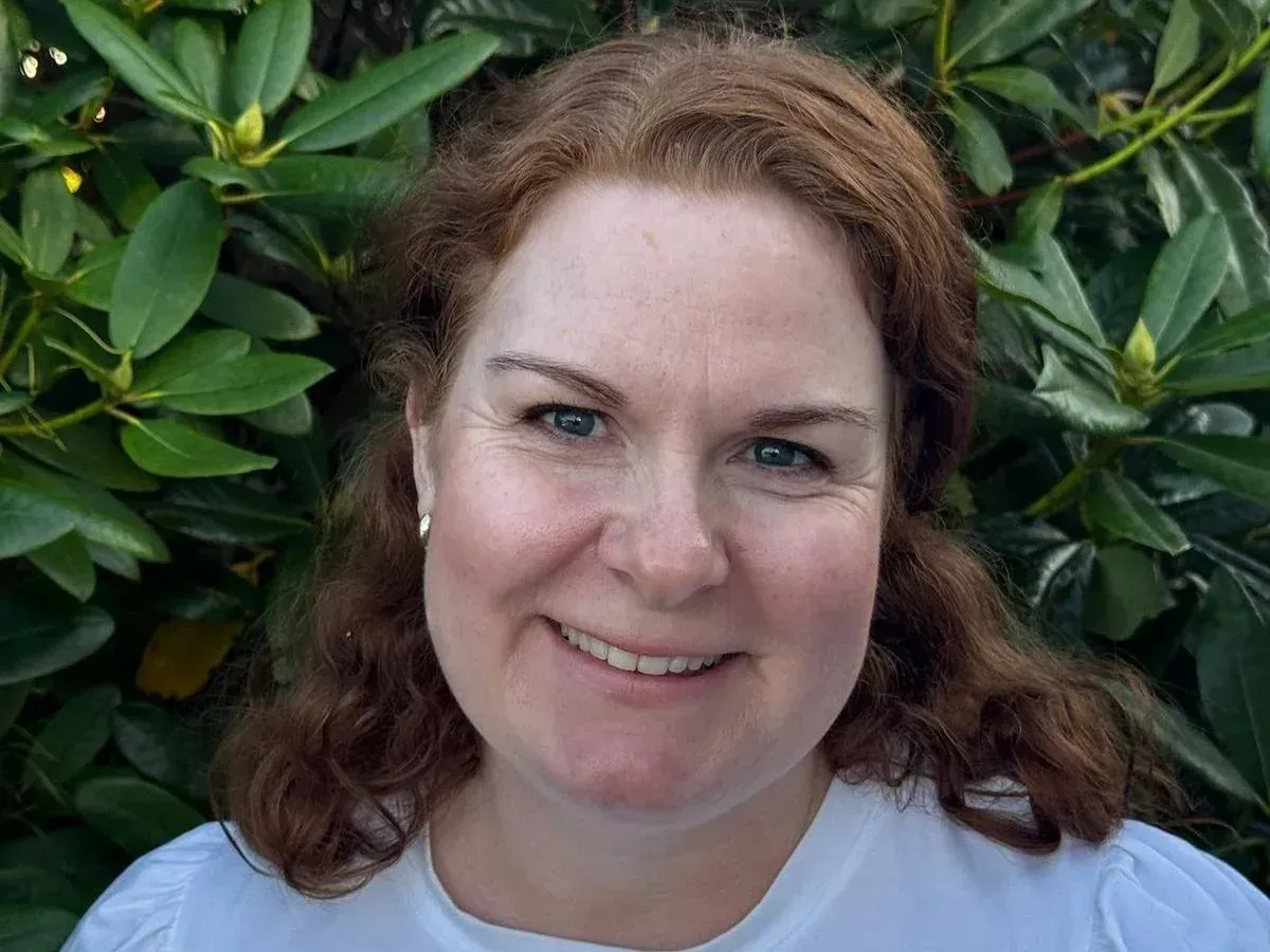 En person med bølget rødbrunt hår som smiler mykt foran en løvgrønn busk.