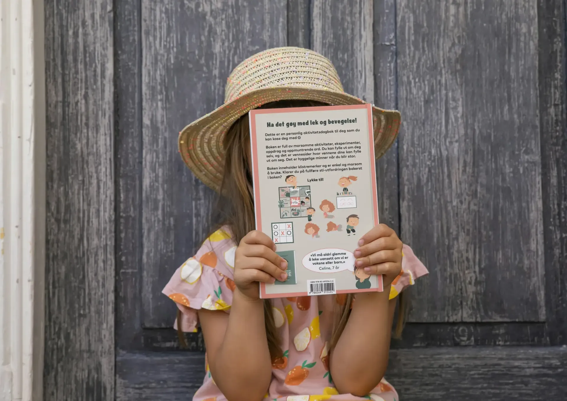 En person med stråhatt holder en lys bok foran ansiktet sitt, sittende foran en grå tredør.