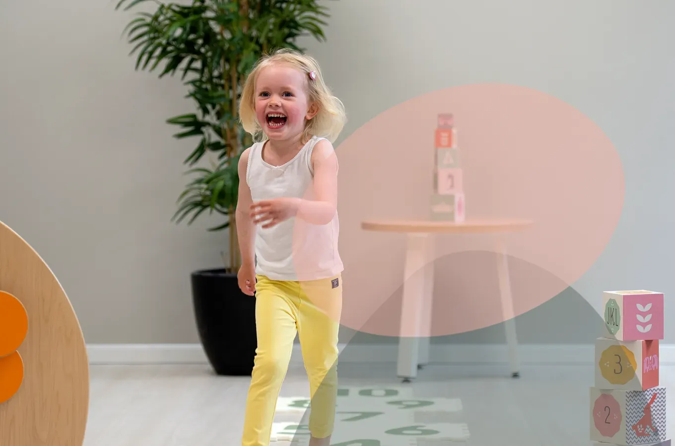 Et gledesfylt barn i en hvit singlet og gule bukser løper over et lyst innendørsrom i nærheten av en plante og stablede klosser.