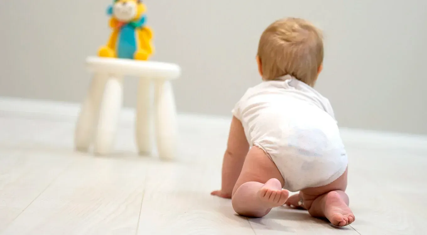 En baby i en hvit bleie kryper på et lyst gulv mot et fargerikt kosedyr som sitter på en liten hvit krakk.