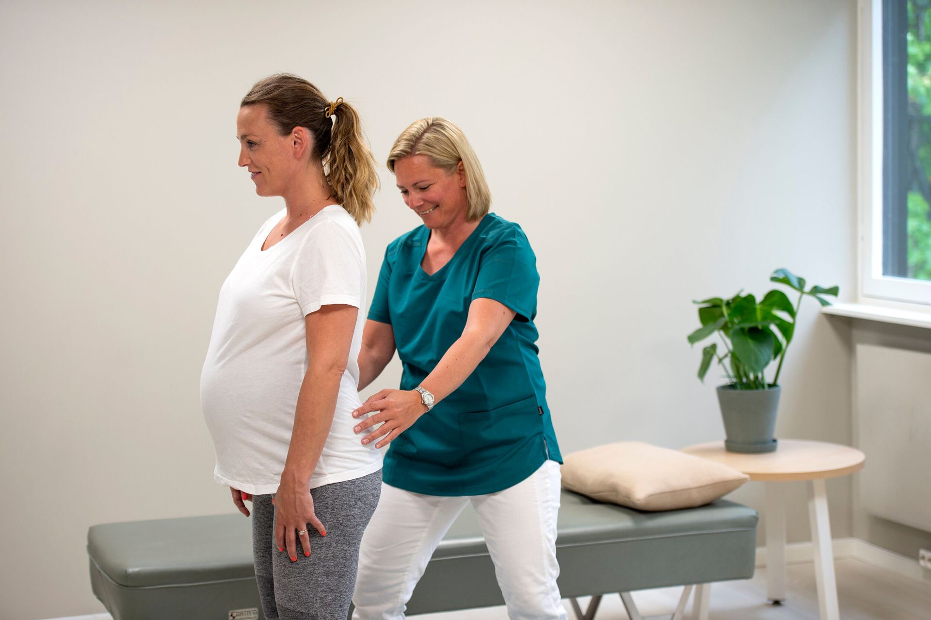 En fysioterapeut står bak en gravid pasient og veileder henne med hendene på ryggen i et klinisk kontormiljø.