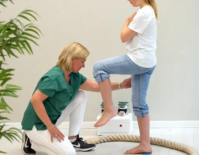 En fysioterapeut i grønne skrubber vurderer en pasients balanse mens de står på ett ben i en klinikksetting.