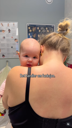 En person holder en baby mot skulderen sin i et rom med medisinske plakater på veggen. Teksten sier «Behandler en fostep».