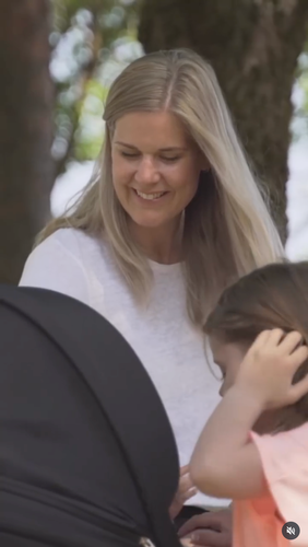 En person smiler mens hun ser ned på en barnevogn ved siden av et barn utendørs.