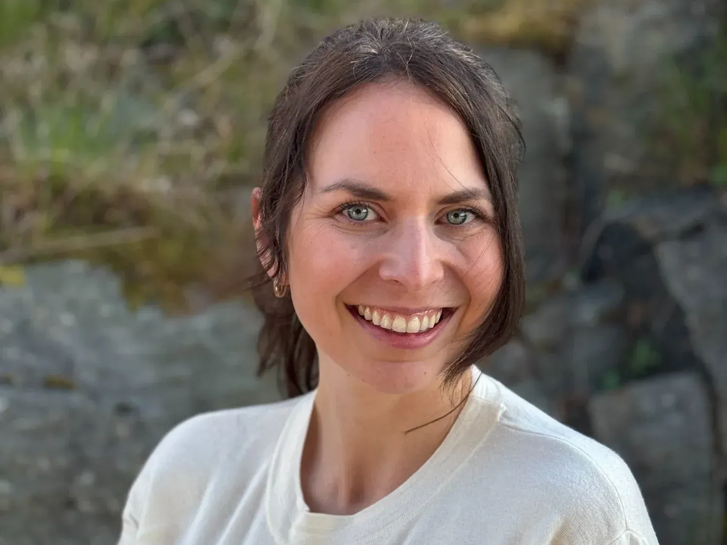En smilende person iført hvit skjorte, plassert utendørs med en uskarp, naturlig bakgrunn.