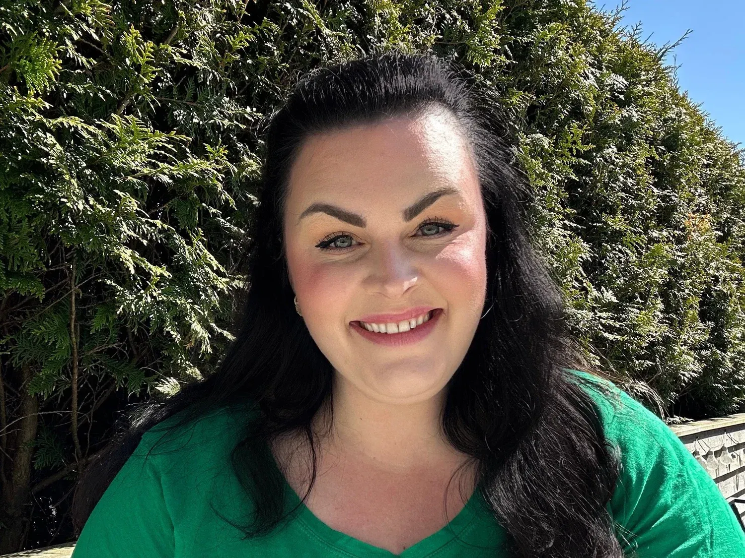 En smilende person med mørkt hår iført en grønn skjorte, plassert foran en løvgrønn hekk på en solrik dag.