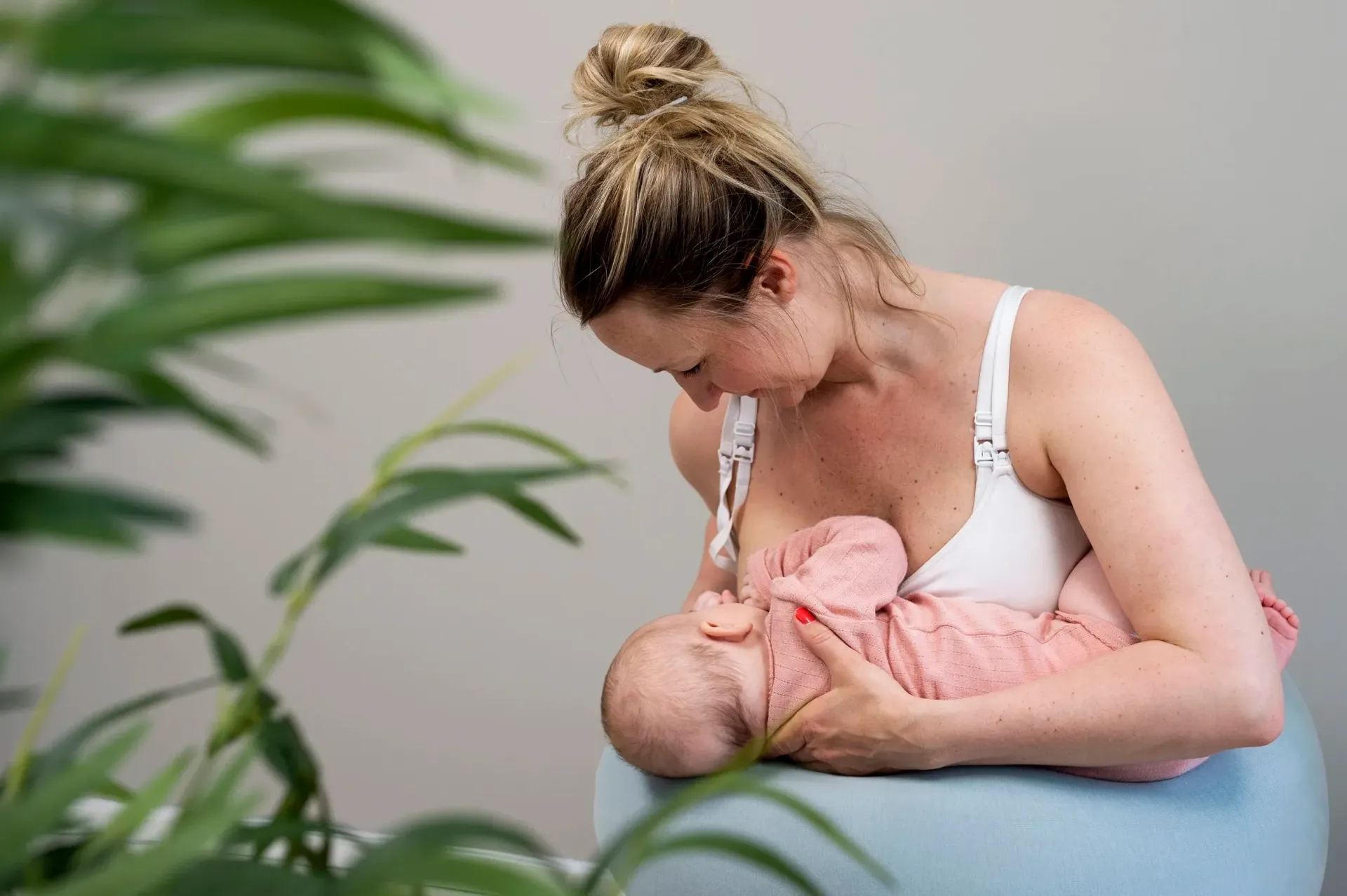 En person ammer en nyfødt mens hun sitter på en lyseblå treningsball, innrammet av grønne blader i forgrunnen.