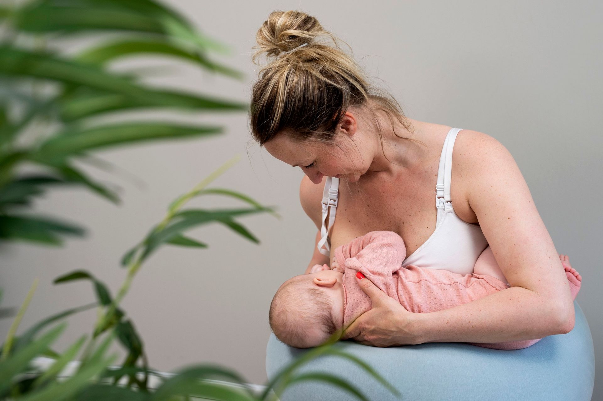 En person som sitter på en blå pute og ammer en baby, innrammet av grønne blader i forgrunnen.