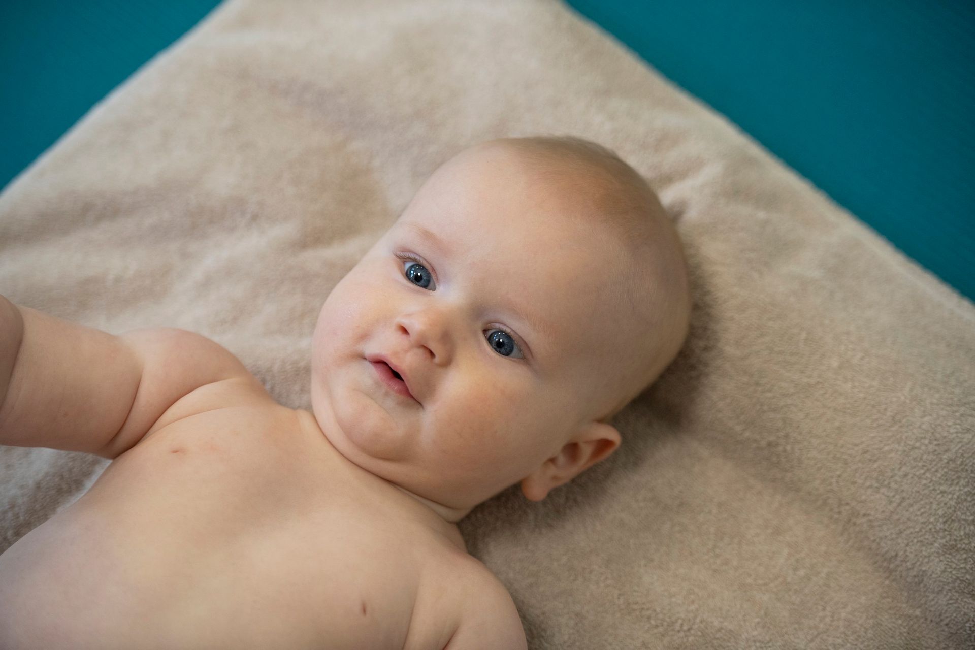 En lyshudet baby med blå øyne som ligger på et brunt teppe mot en blågrønn bakgrunn.