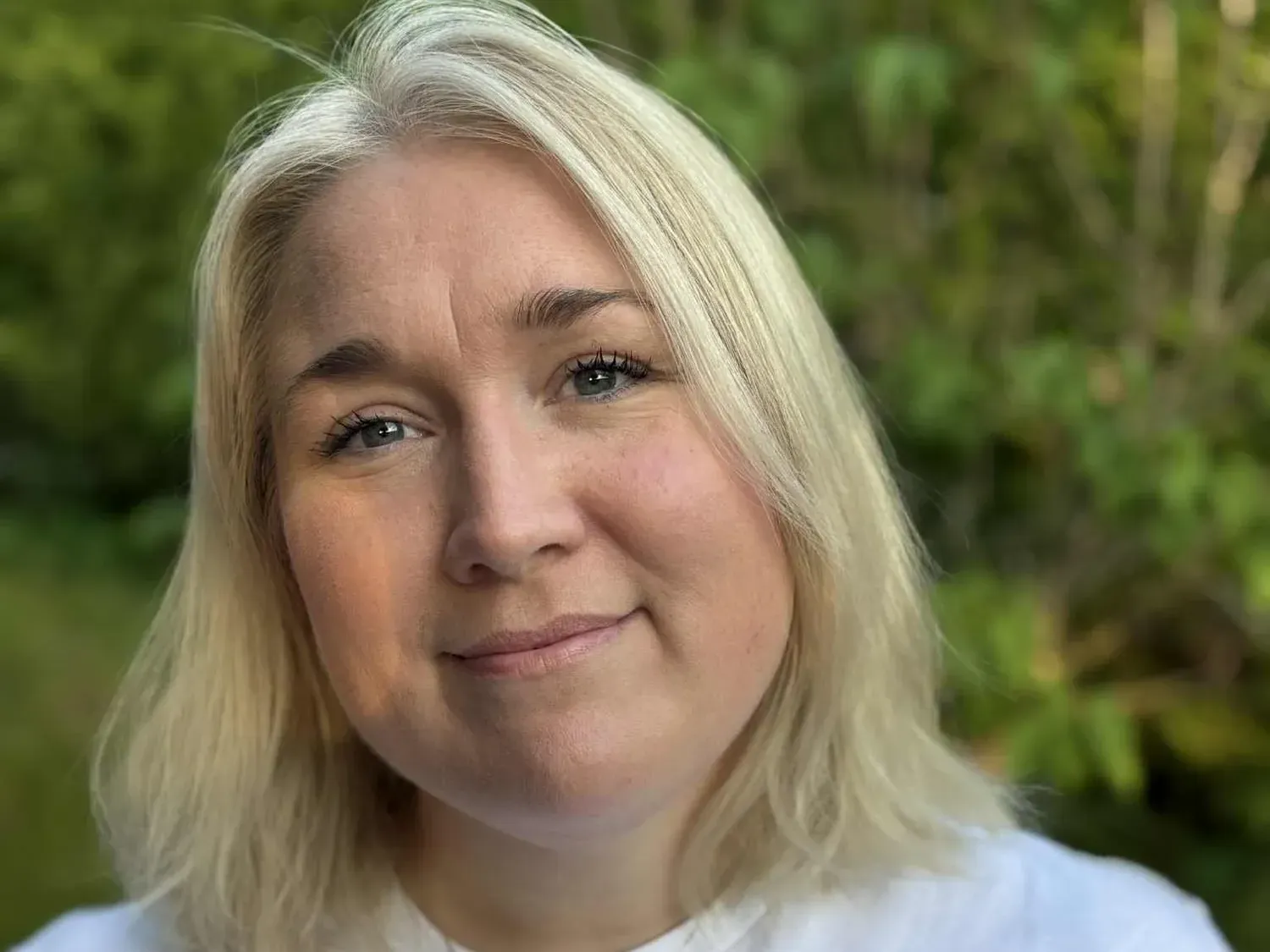 En person med skulderlangt blondt hår poserer foran en uskarp grønn, naturlig bakgrunn.