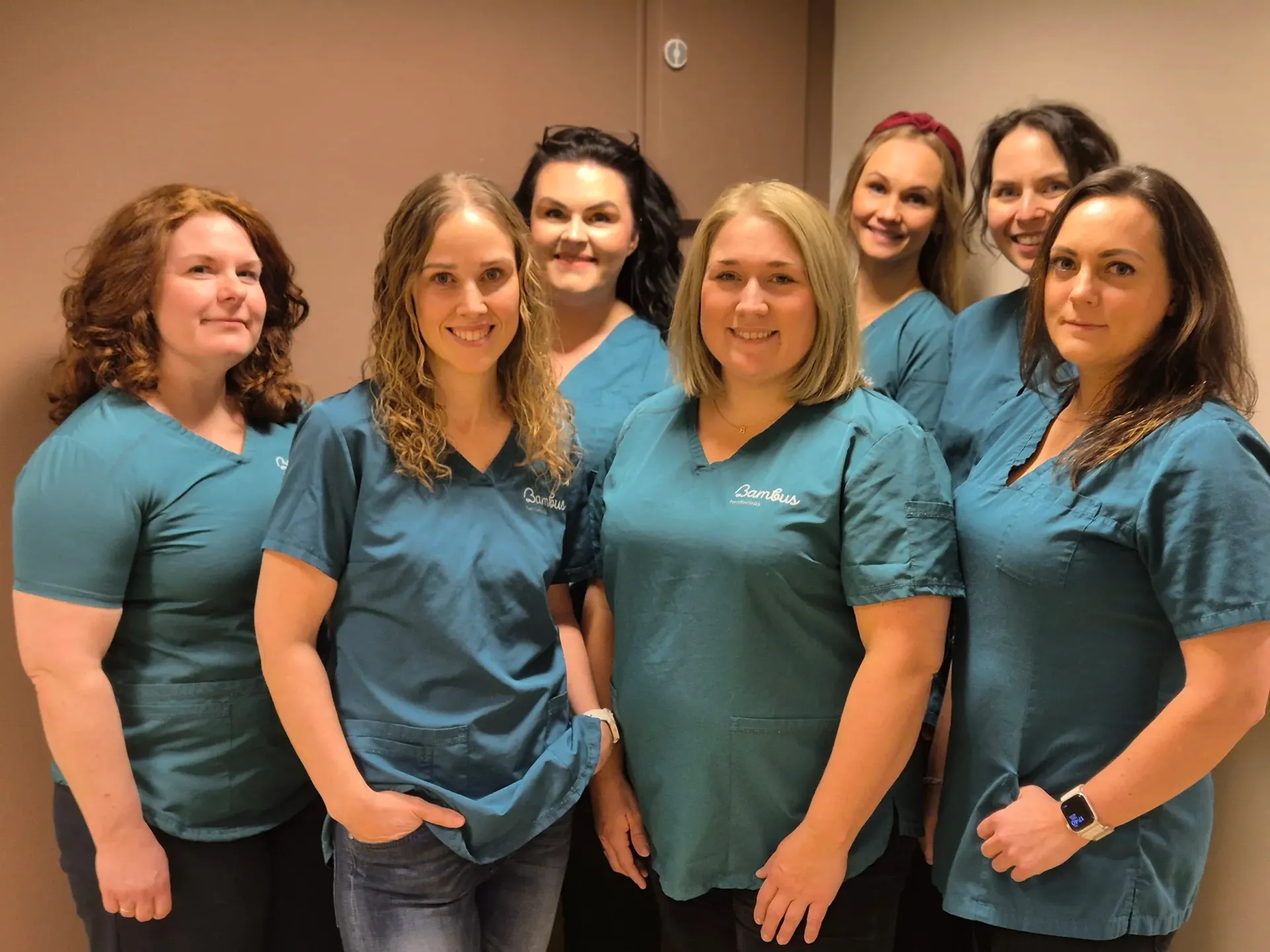 Syv personer står i en kø mot en nøytral vegg, smilende og har på seg matchende blågrønne skrubber.