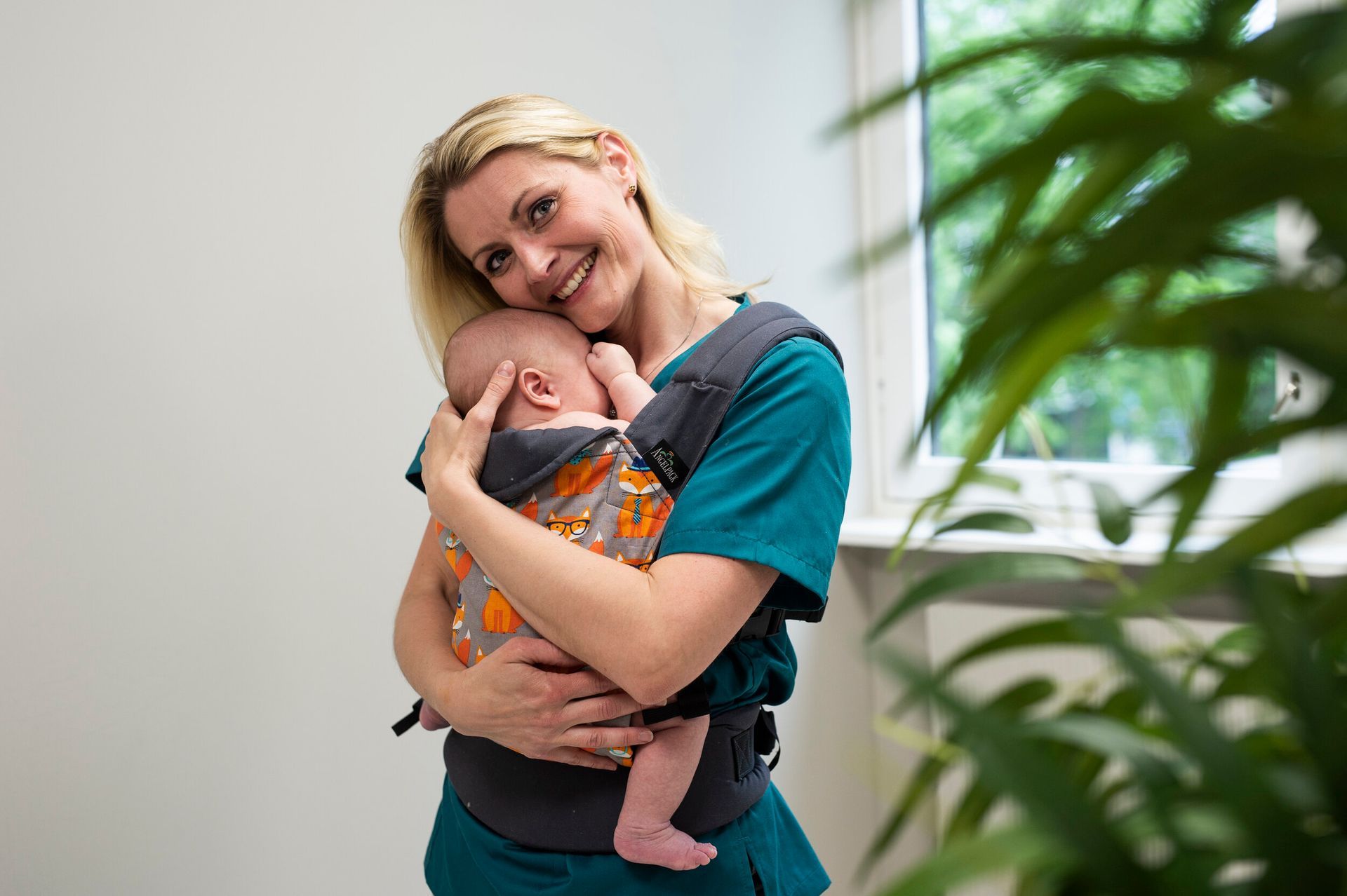 En smilende person i en grønn skjorte har på seg en grå bæresele med et oransje revemønster og holder et sovende spedbarn.