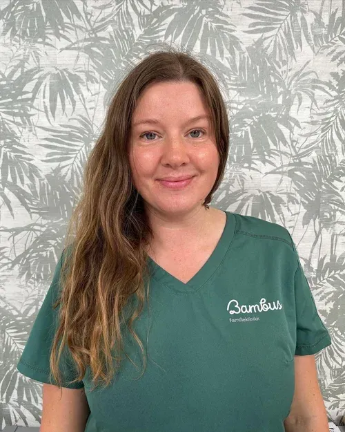 En person med langt brunt hår iført en grønn «Bambus»-uniformtopp, poserer foran en palmebladmønstret bakgrunn.