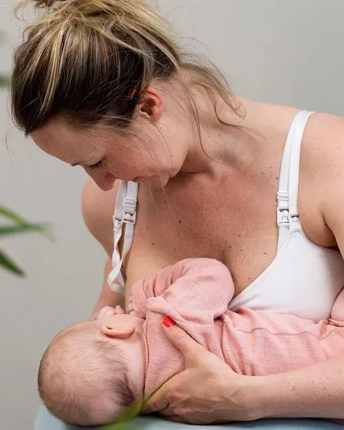 En person i hvit BH vugger og ammer en baby kledd i rosa bodystocking.