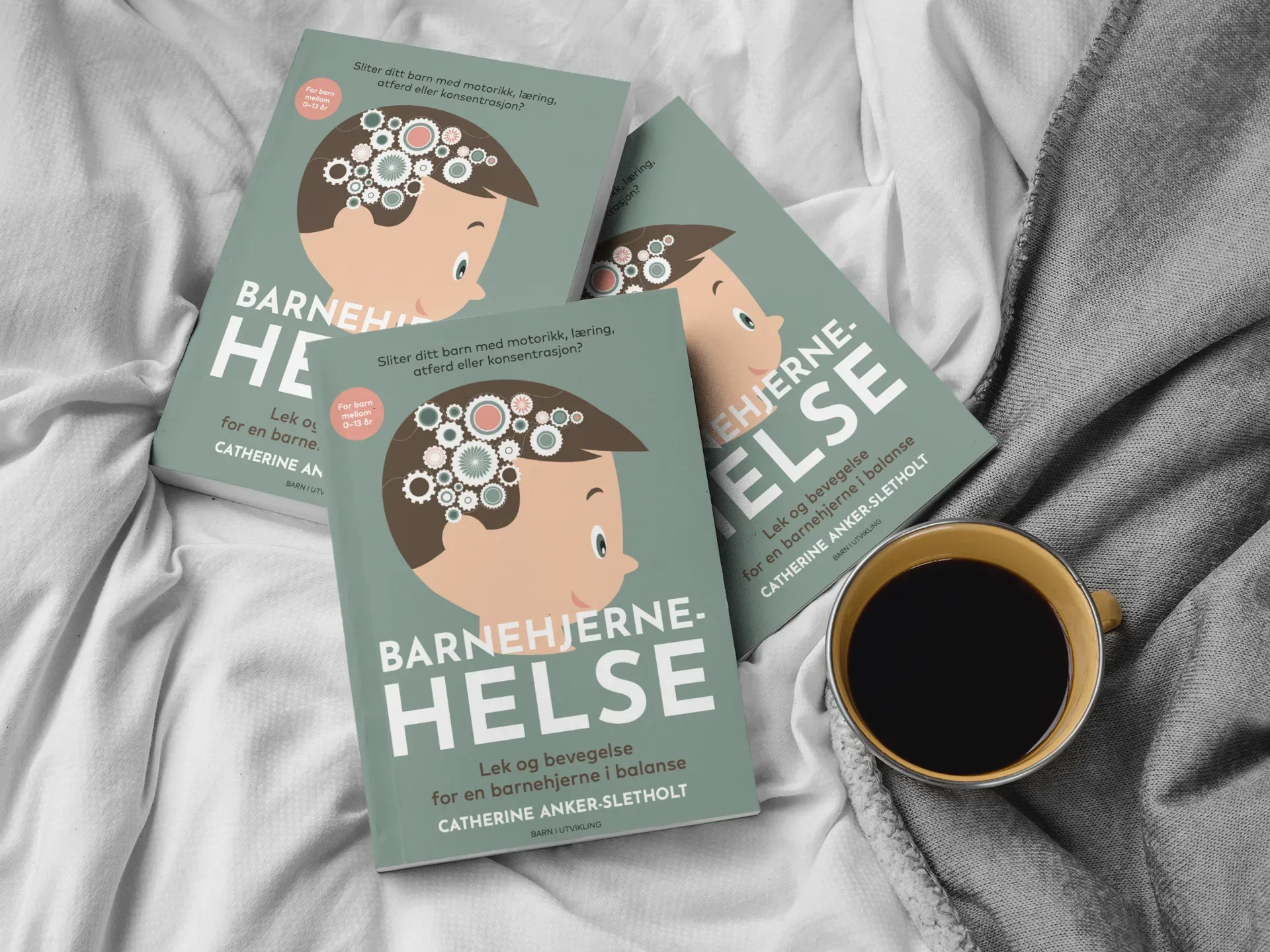 Tre eksemplarer av boken «Barnehjernehelse» arrangert på et hvitt ark ved siden av en kopp svart kaffe.