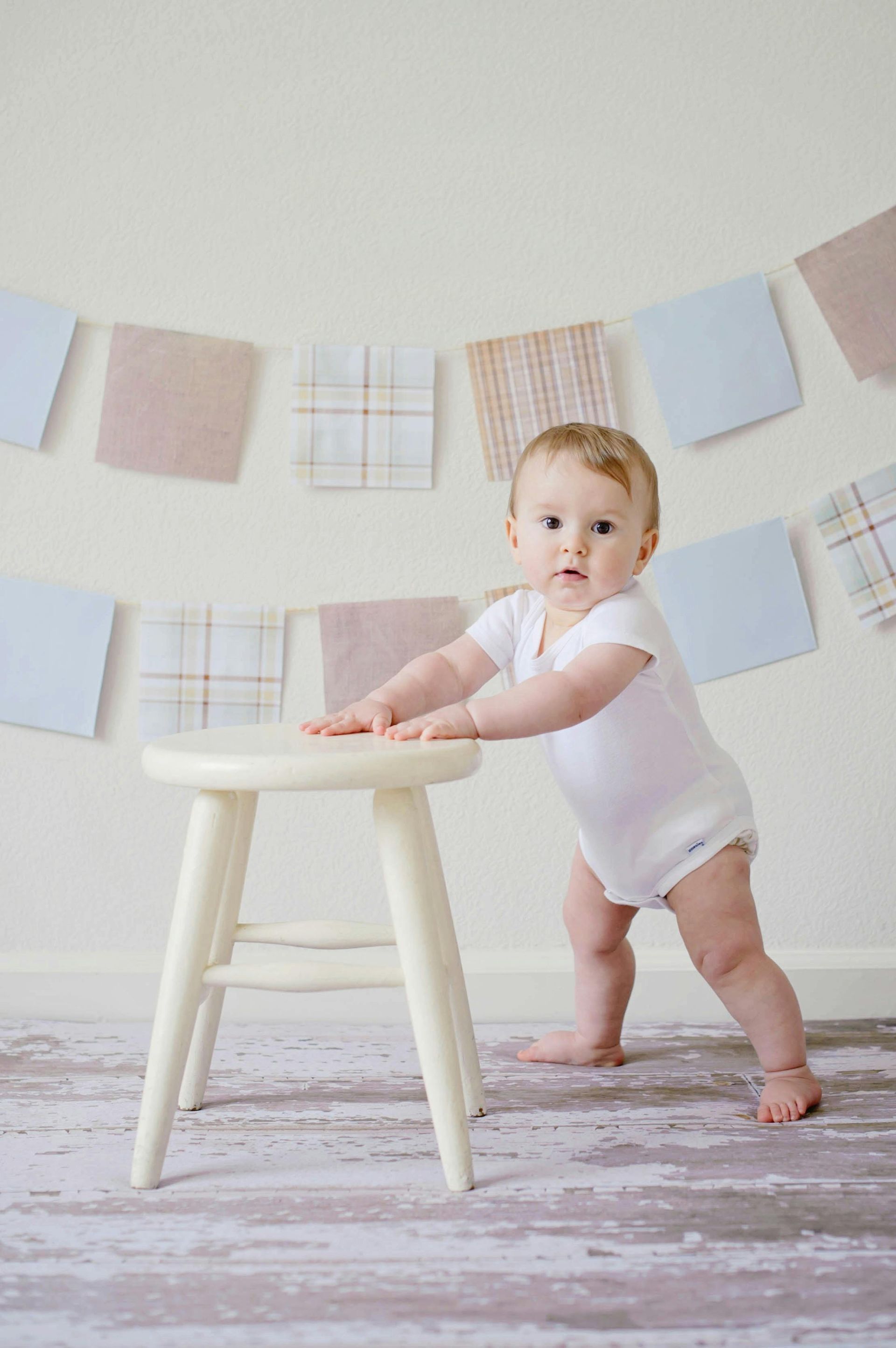 En baby som står og holder seg fast i en hvit trekrakk, med et dekorativt banner av stoffkvadrater i bakgrunnen.