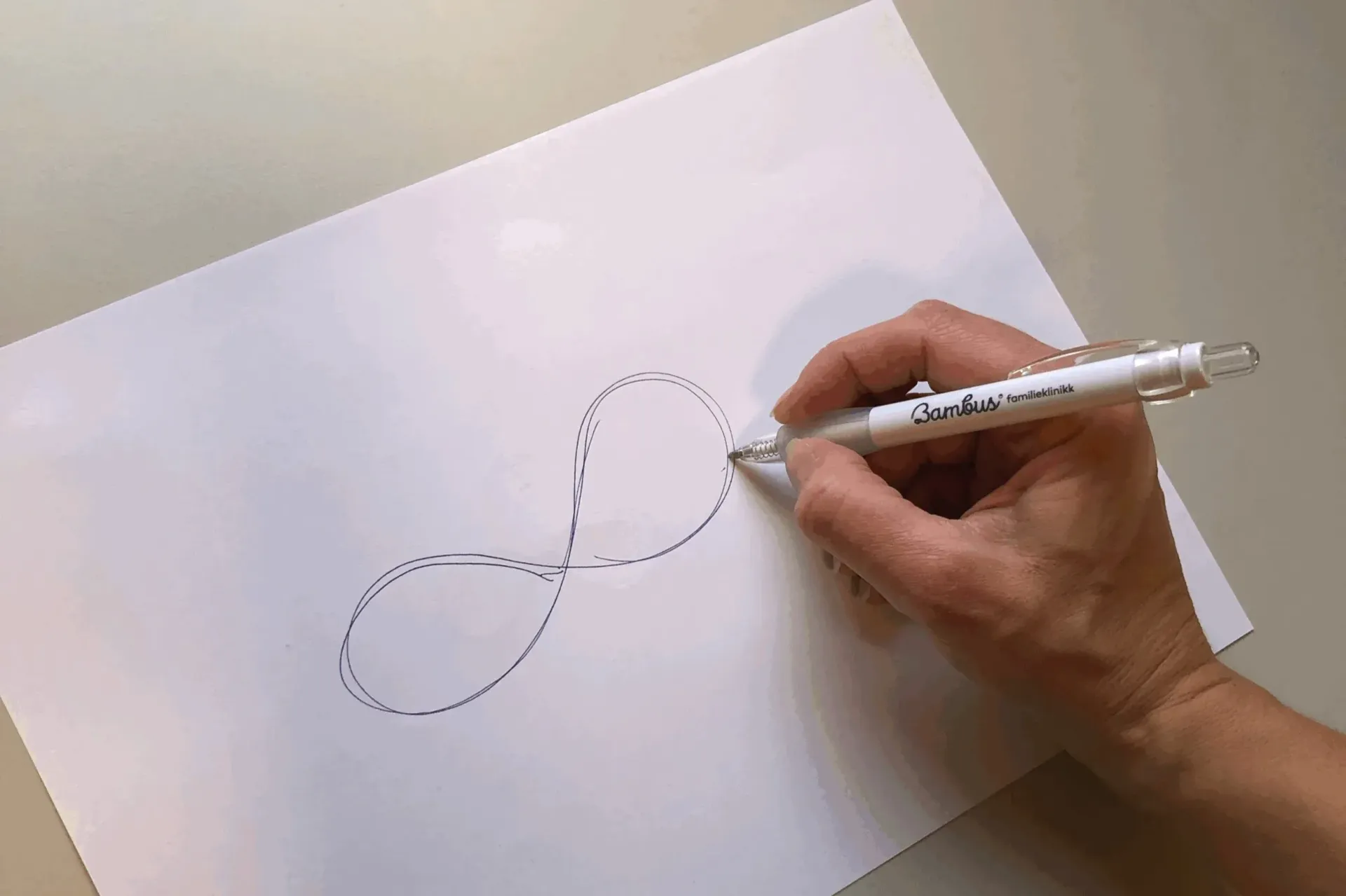 En hånd bruker en penn til å tegne et uendelighetssymbol på et hvitt ark.