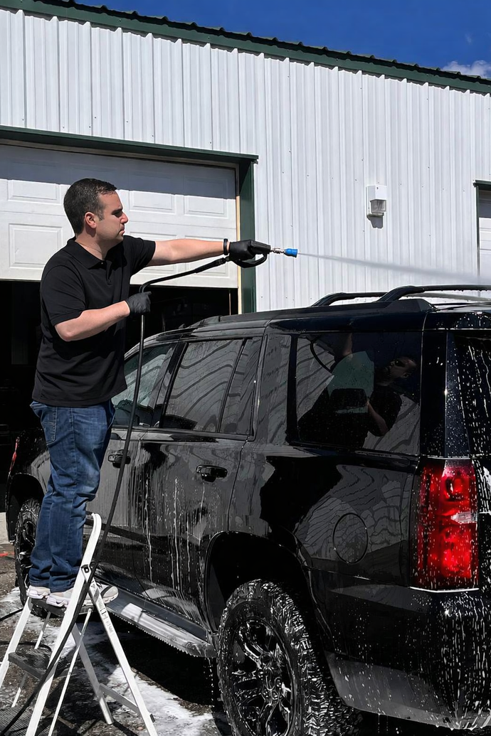 Man washing a black SUV with a pressure washer outside a white building