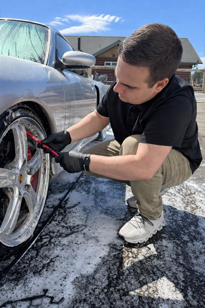 Man cleaning a silver car wheel with a red brush in a sunny driveway