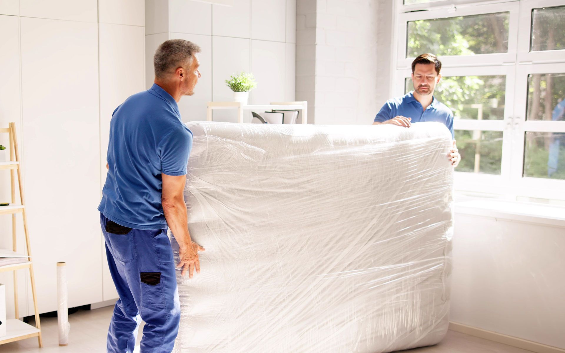 Two men are carrying a large mattress wrapped in plastic.