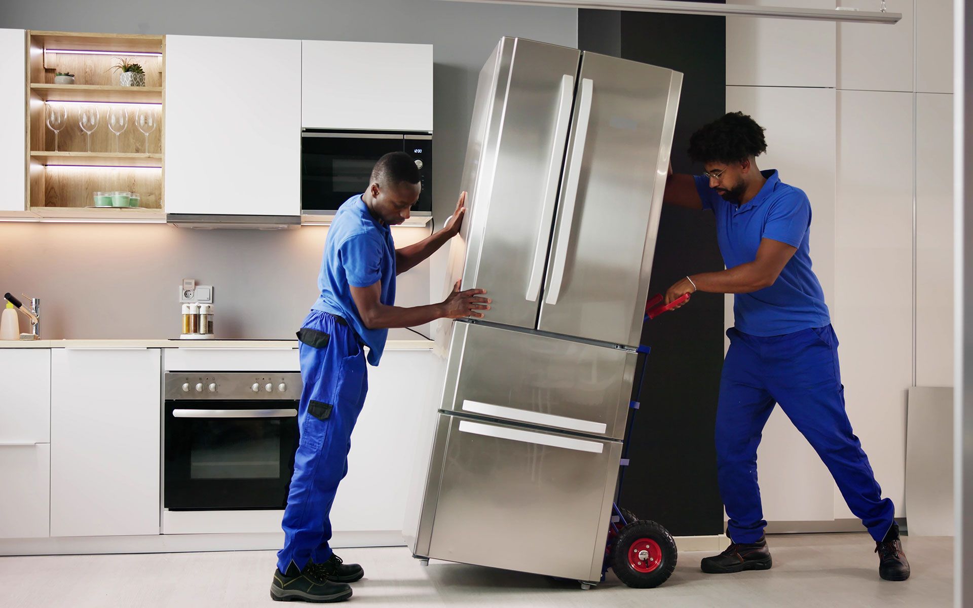 Two men are carrying a refrigerator on a dolly in a kitchen.