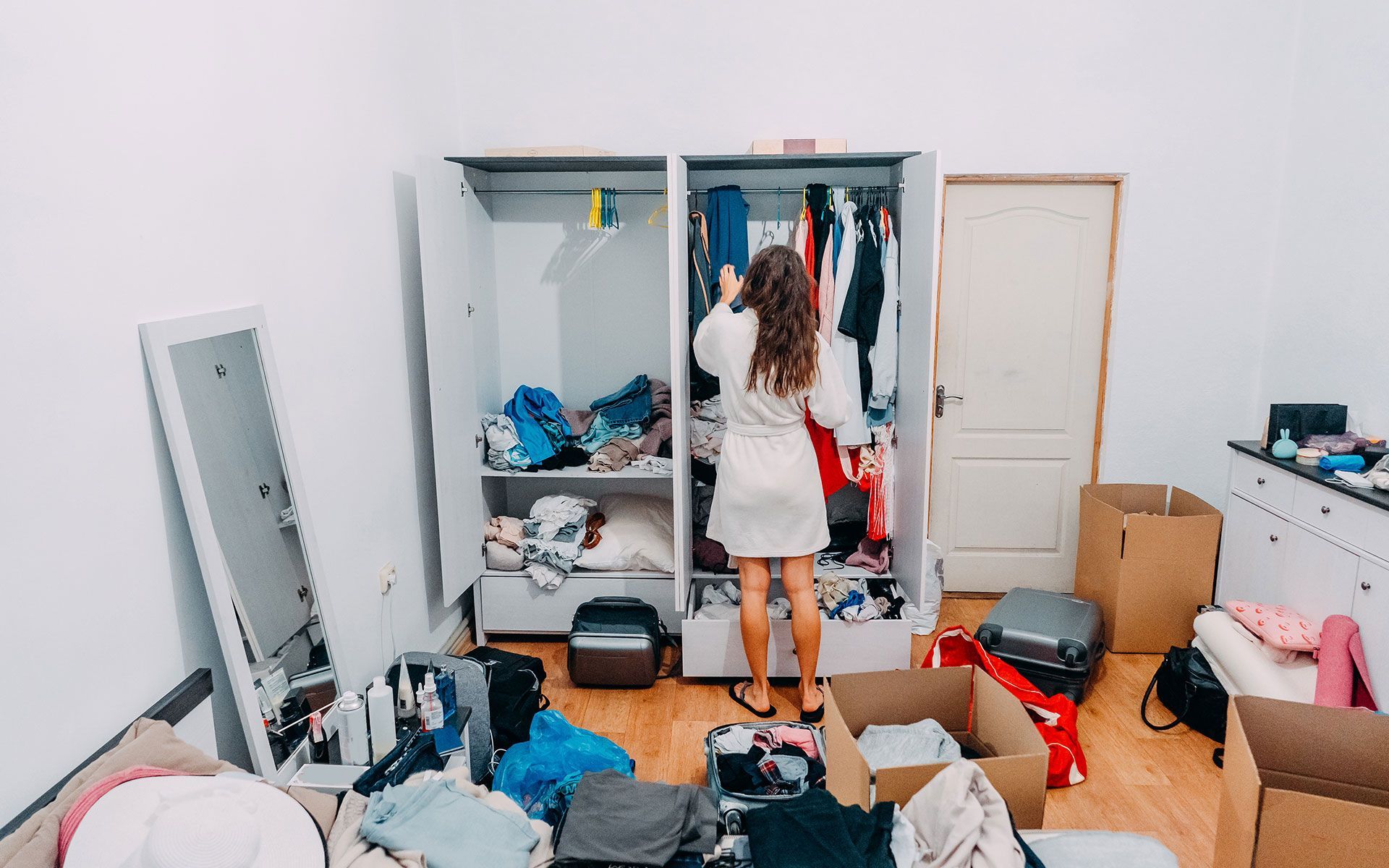 A woman is standing in a messy room looking in a closet.