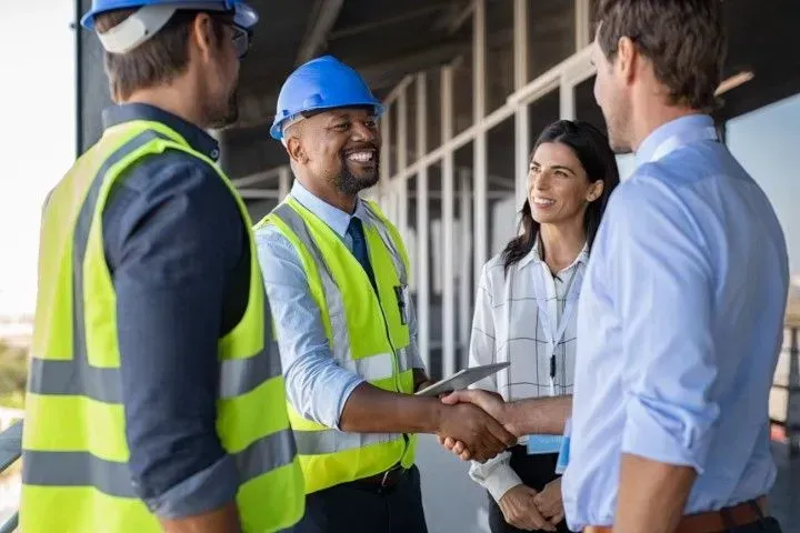 Workers in hard hats and safety vests shake hands at a construction site, smiling and talking.