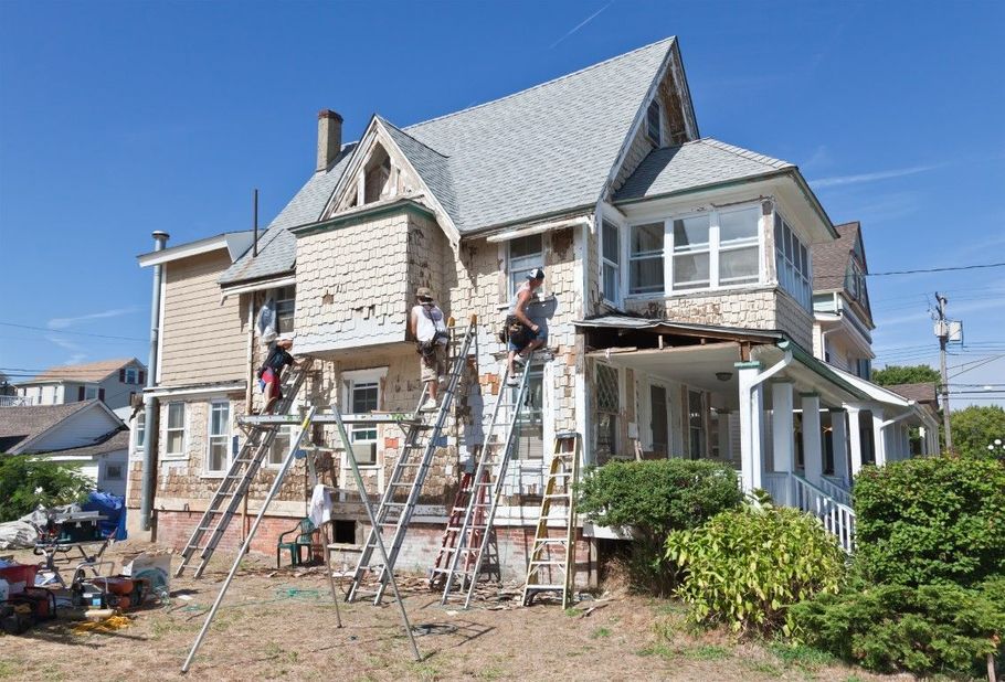 Workers on ladders painting and repairing a two-story house exterior with scaffolding.