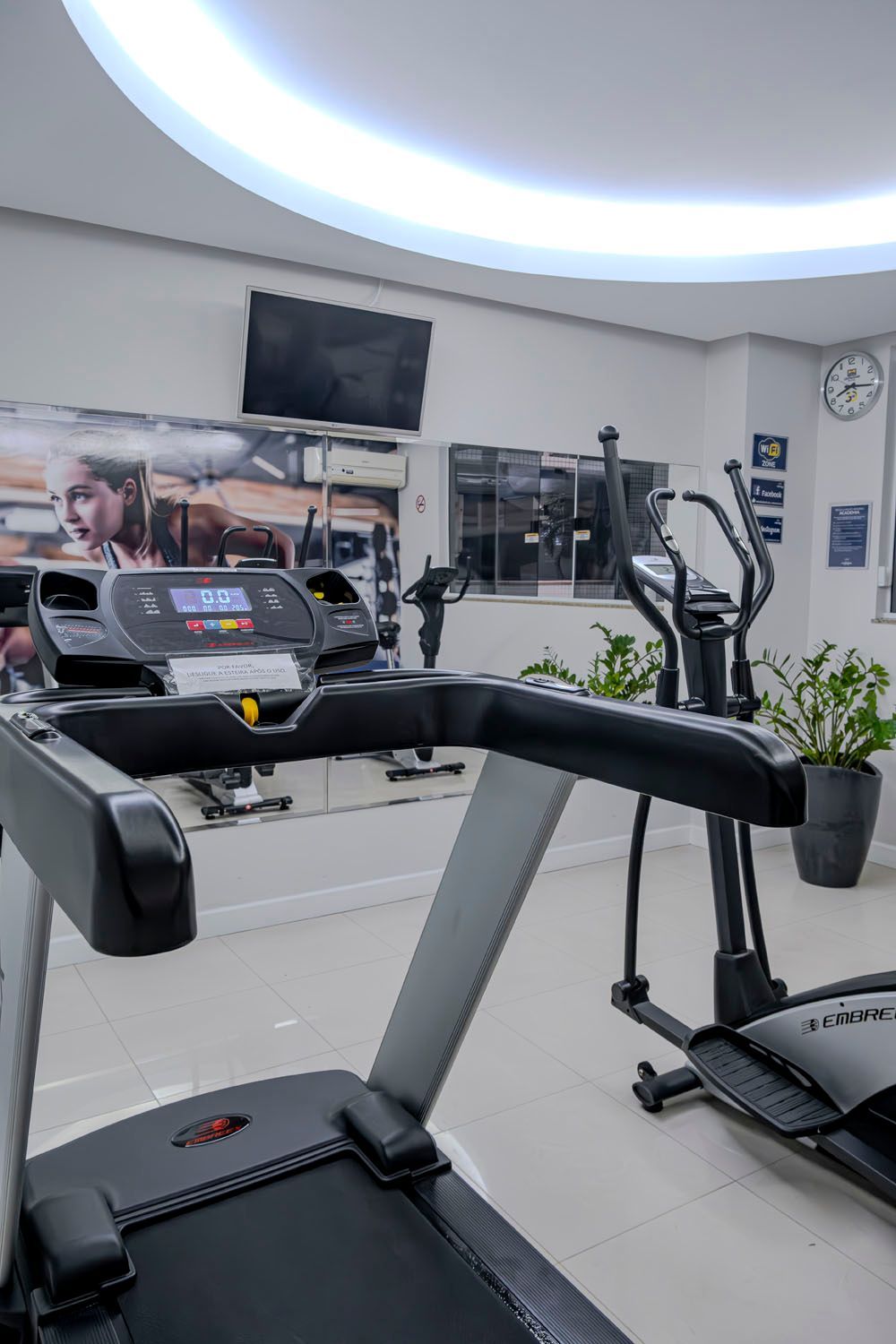 Uma academia com esteiras, elípticos e uma televisão.