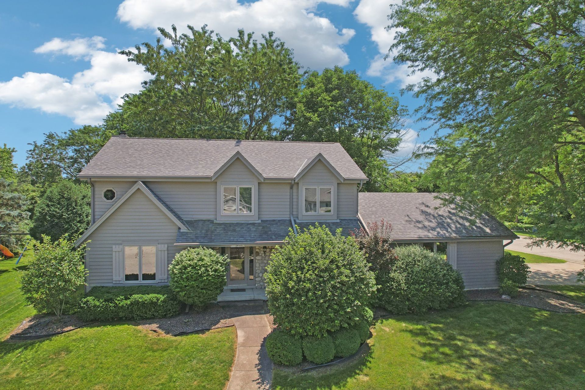 Gray two-story house with green lawn and trees under a blue sky.