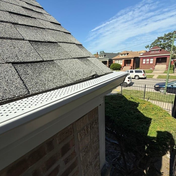 Roof with grey shingles and white gutter with guard, houses in background. Sunny day.