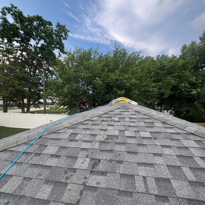 Roofer on gray shingle roof, blue safety rope, trees, white fence, cloudy sky.