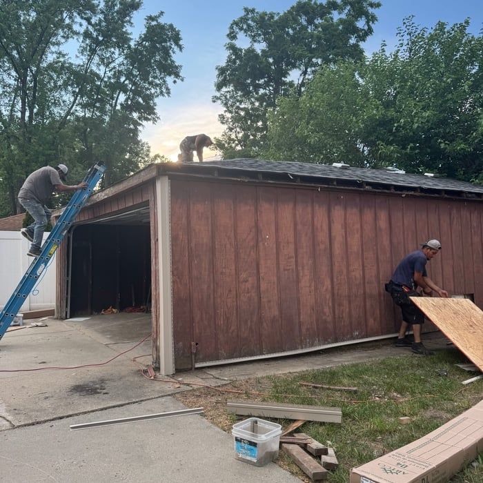 Workers on a roof, cutting and installing wood siding. Garage exterior with ladder, tools, and trees.